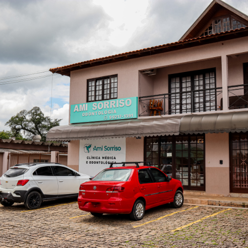 Fachada da clínica Ami Sorriso em Campo Magro, com placa visível e estacionamento com veículos em frente ao prédio.
