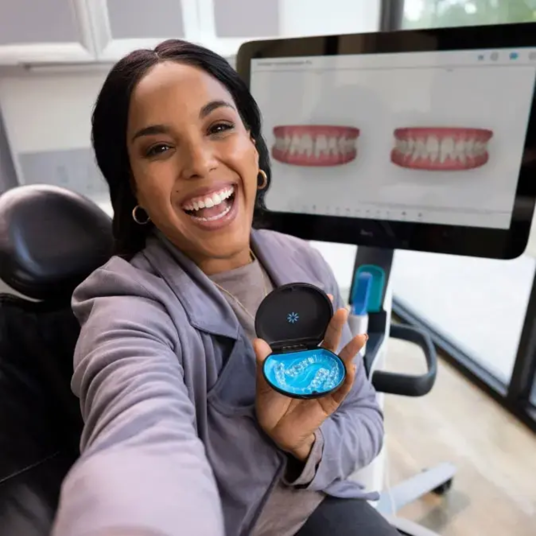 Paciente sorrindo em consultório odontológico enquanto segura o estojo com alinhadores, com imagem digital do sorriso ao fundo.