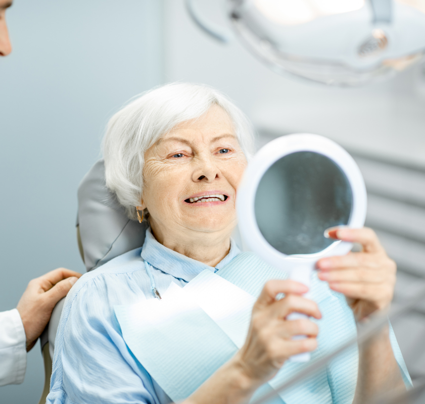 Idosa sorrindo ao se ver no espelho após procedimento odontológico, representando autoestima recuperada com implantes dentários